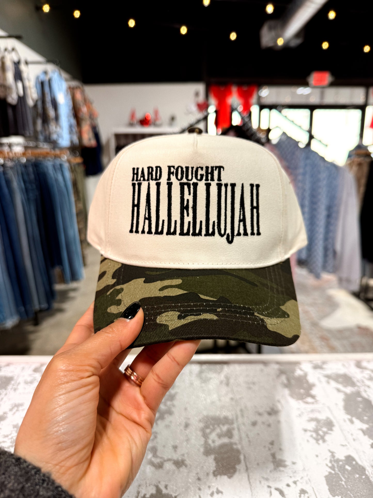 Camo Cap with 'Hard Fought Hallelujah' text held in a store setting