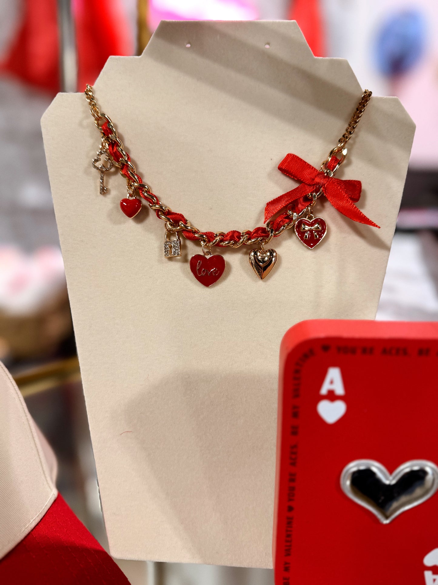 Necklace with red heart charms and a red bow on a display stand, with a playing card featuring a heart symbol in the background.