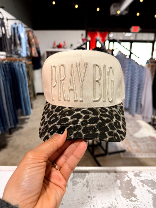 Cap with 'PRAY BIG' text held in front of a store interior.