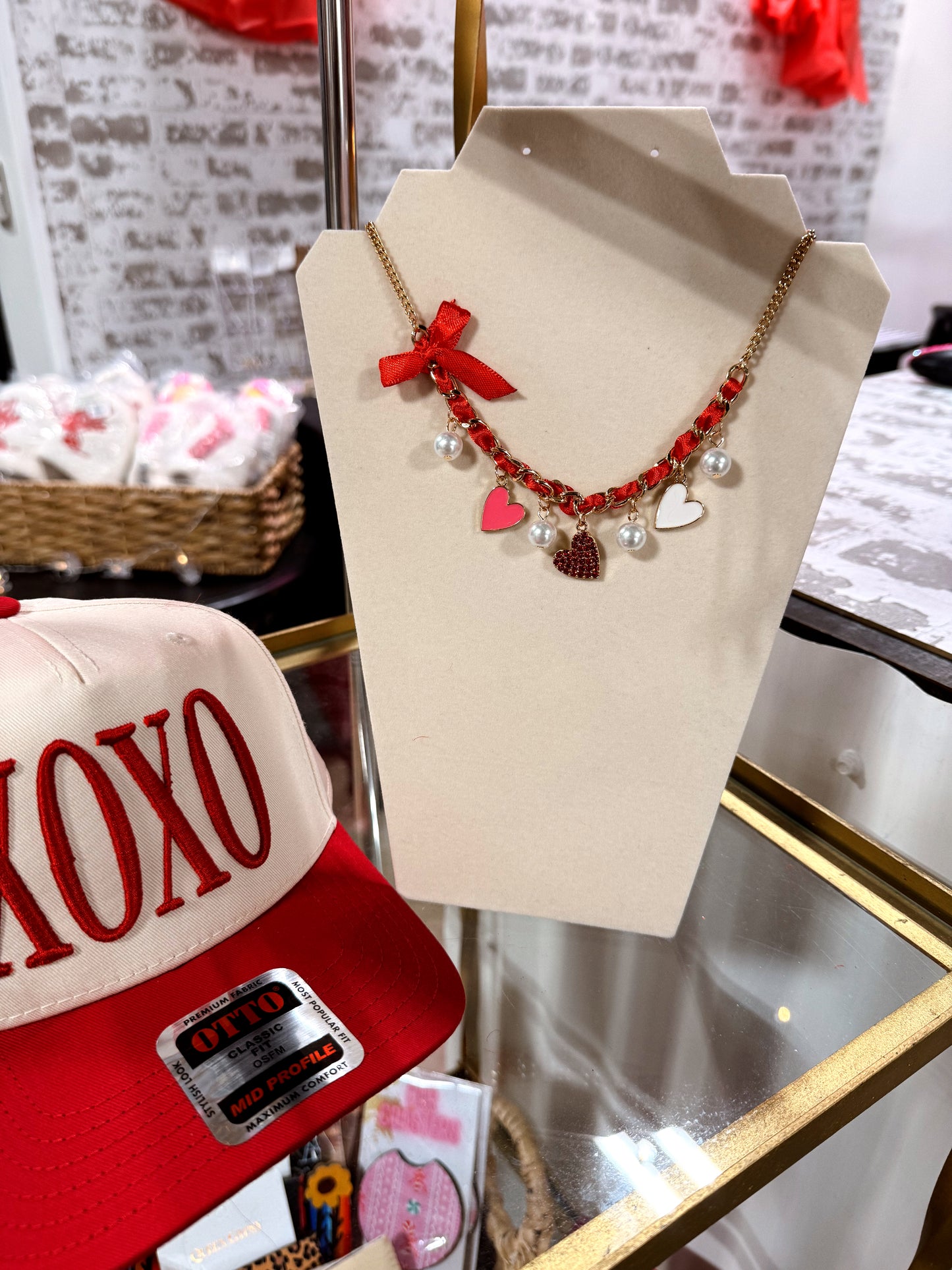 Necklace with red bows and heart charms on a mannequin bust, with a 'XOXO' cap in the foreground.