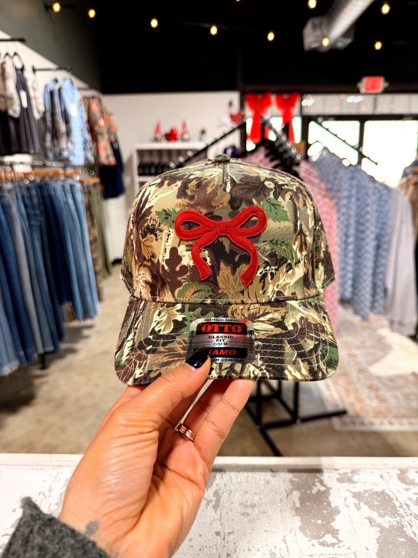 Camouflage cap with a red bow design held in a store setting