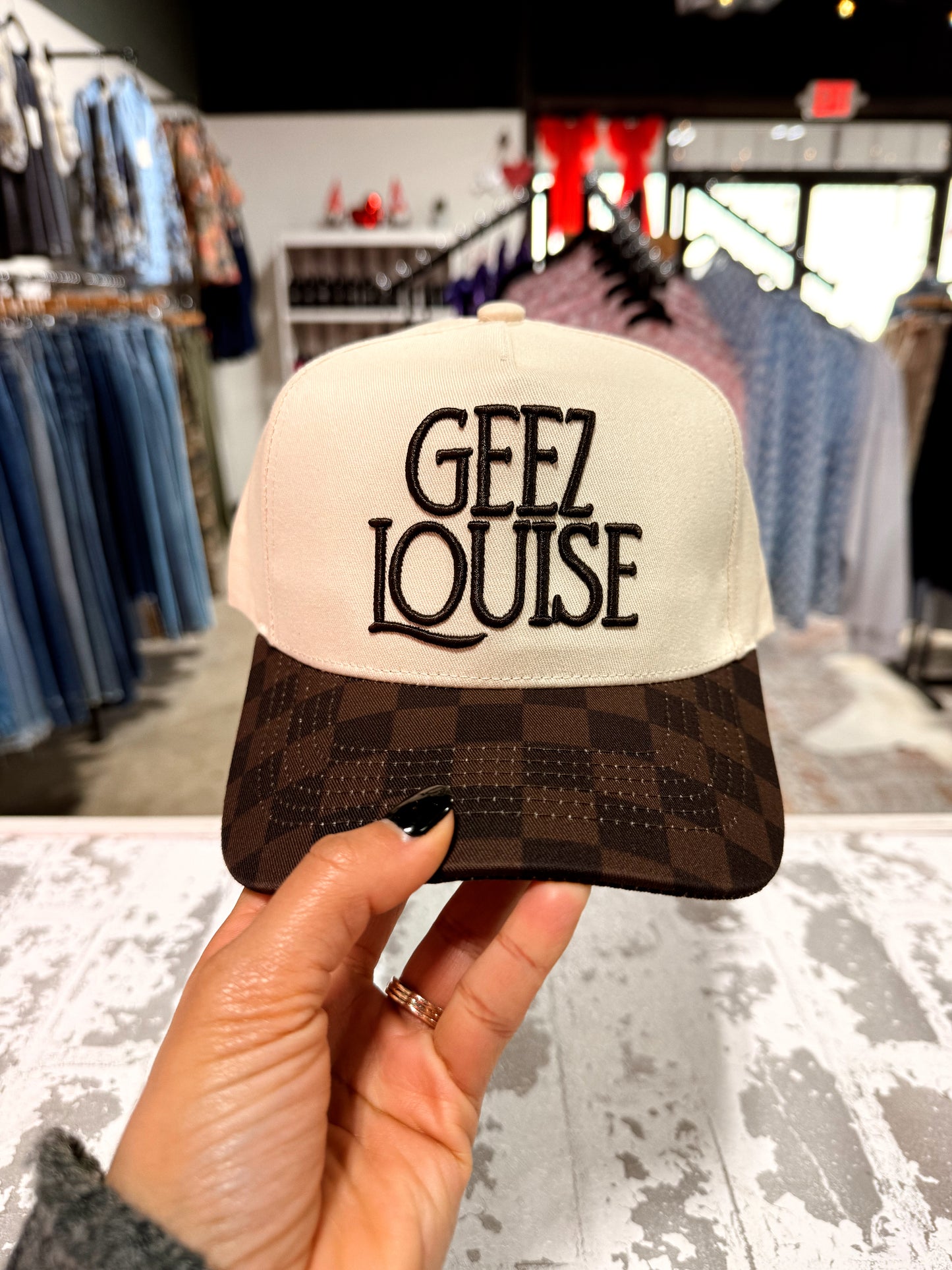 Cap with 'Geez Louise' text held in a hand, with a store interior background.